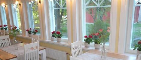 Uma área de refeições com cadeiras e mesas brancas, plantas em vasos com flores vermelhas e grandes janelas com vista para vegetação e um edifício vermelho.