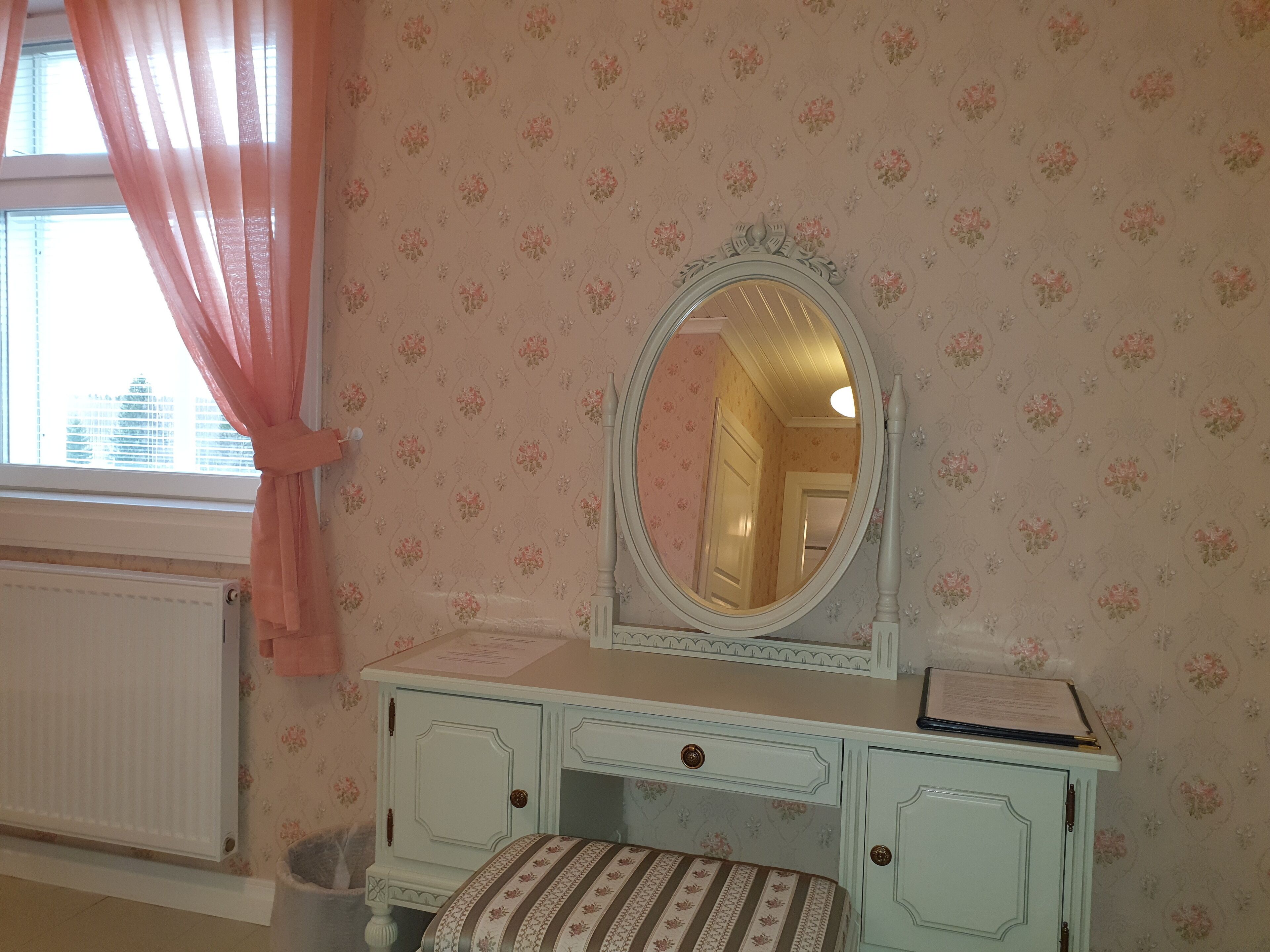 Une pièce comprenant une coiffeuse, un miroir, une fenêtre avec des rideaux roses et un papier peint à motifs floraux.