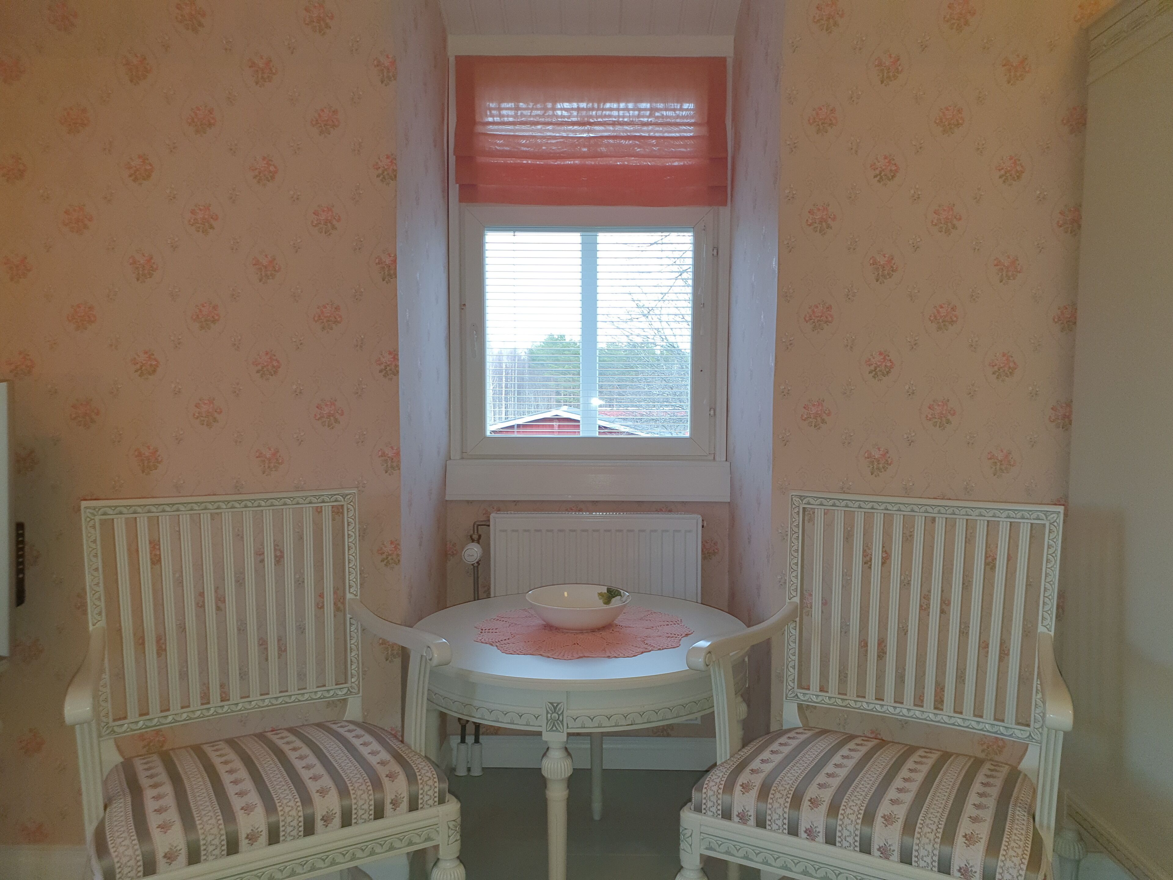 Een kleine kamer met bloemenbehang, een raam met jaloezieën, een radiator en twee gestoffeerde fauteuils met een tafel in het midden.