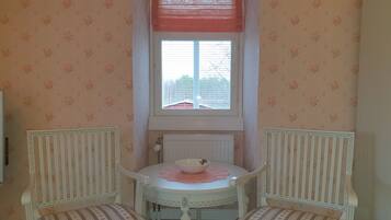 Een kleine kamer met bloemenbehang, een raam met jaloezieën, een radiator en twee gestoffeerde fauteuils met een tafel in het midden.