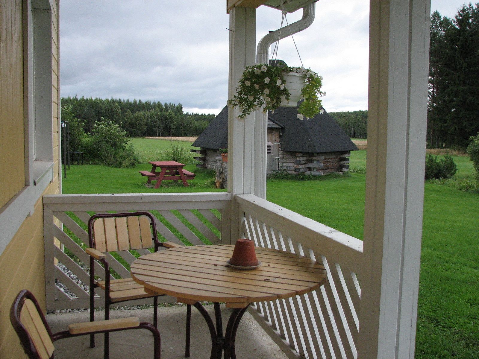 Une véranda avec une table en bois et deux chaises, donnant sur un espace verdoyant où se trouvent un bâtiment en pierre et une table de pique-nique.