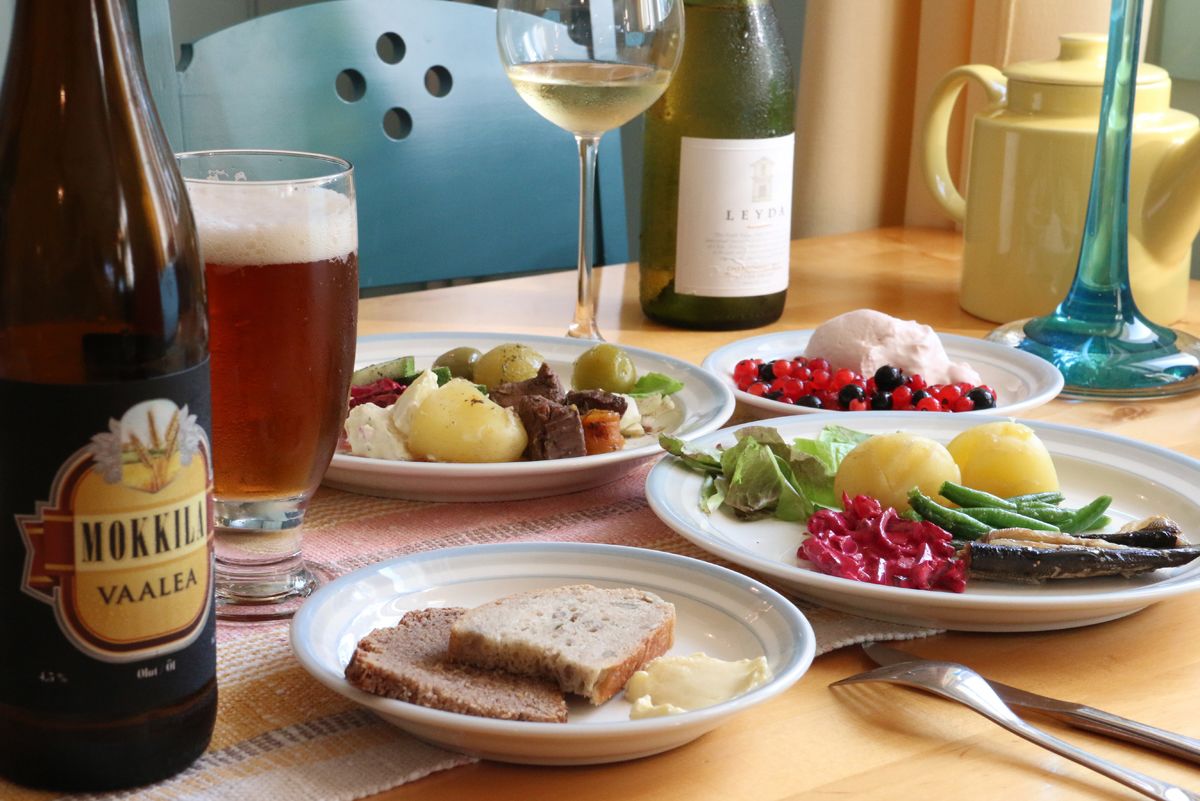 Una mesa con platos de comida, una botella de cerveza Mokkila Vaalea, una copa de vino blanco y una botella de vino Leyla.