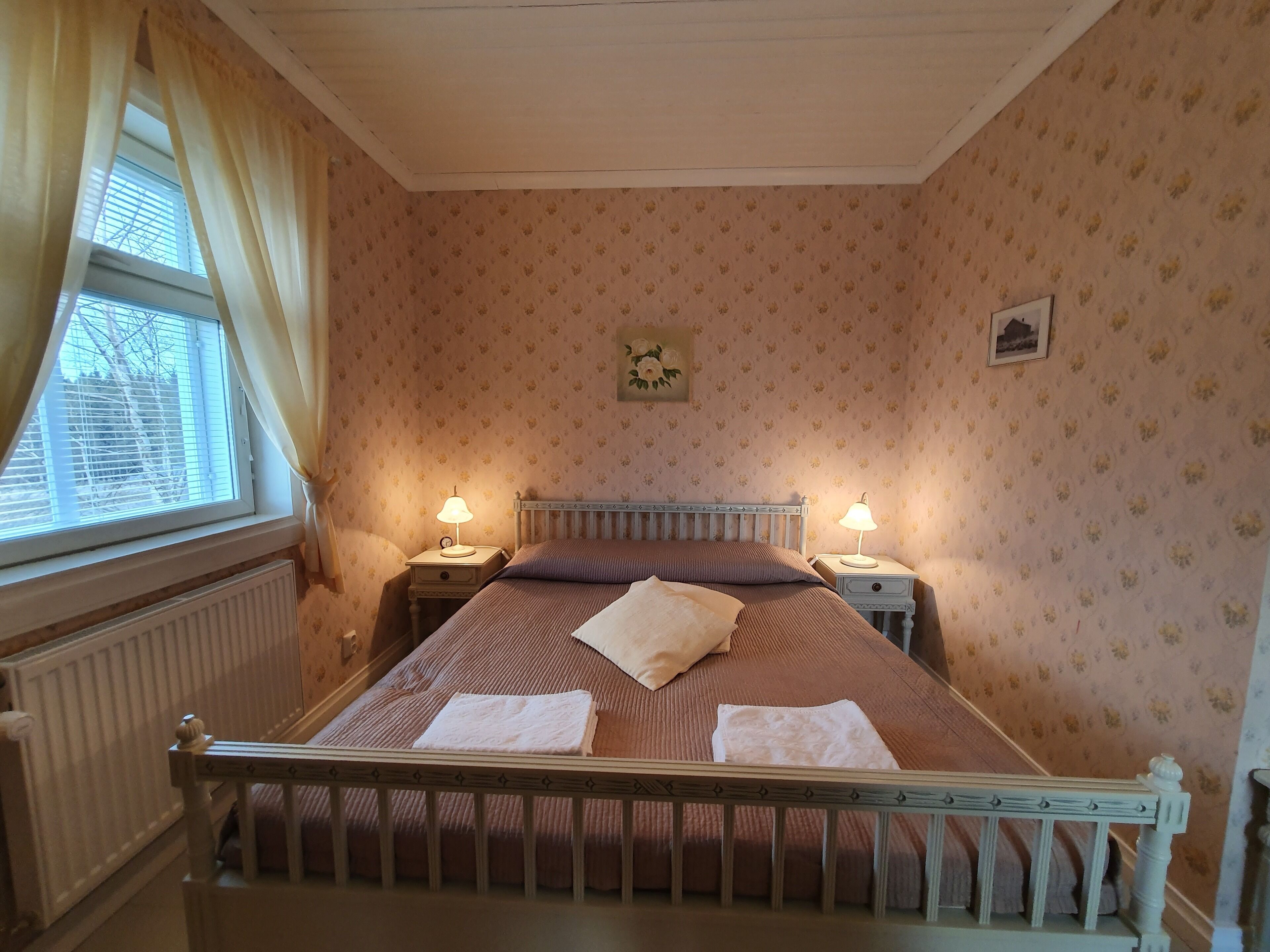 A bedroom with a wooden bed, two bedside tables with lamps, a radiator, a window with curtains, and floral wallpaper.