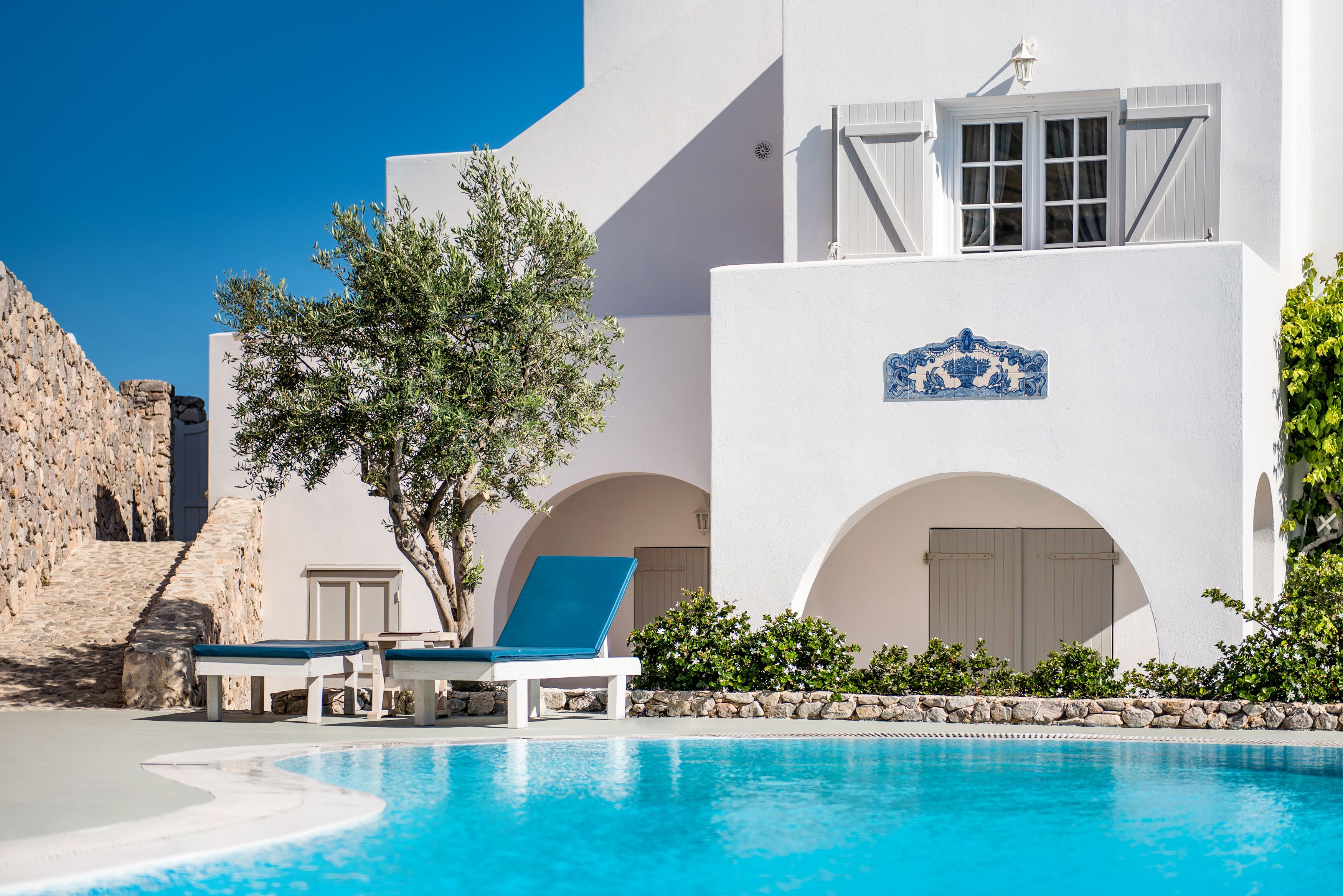 A poolside area with a white building, two blue lounge chairs, and a clear blue pool.