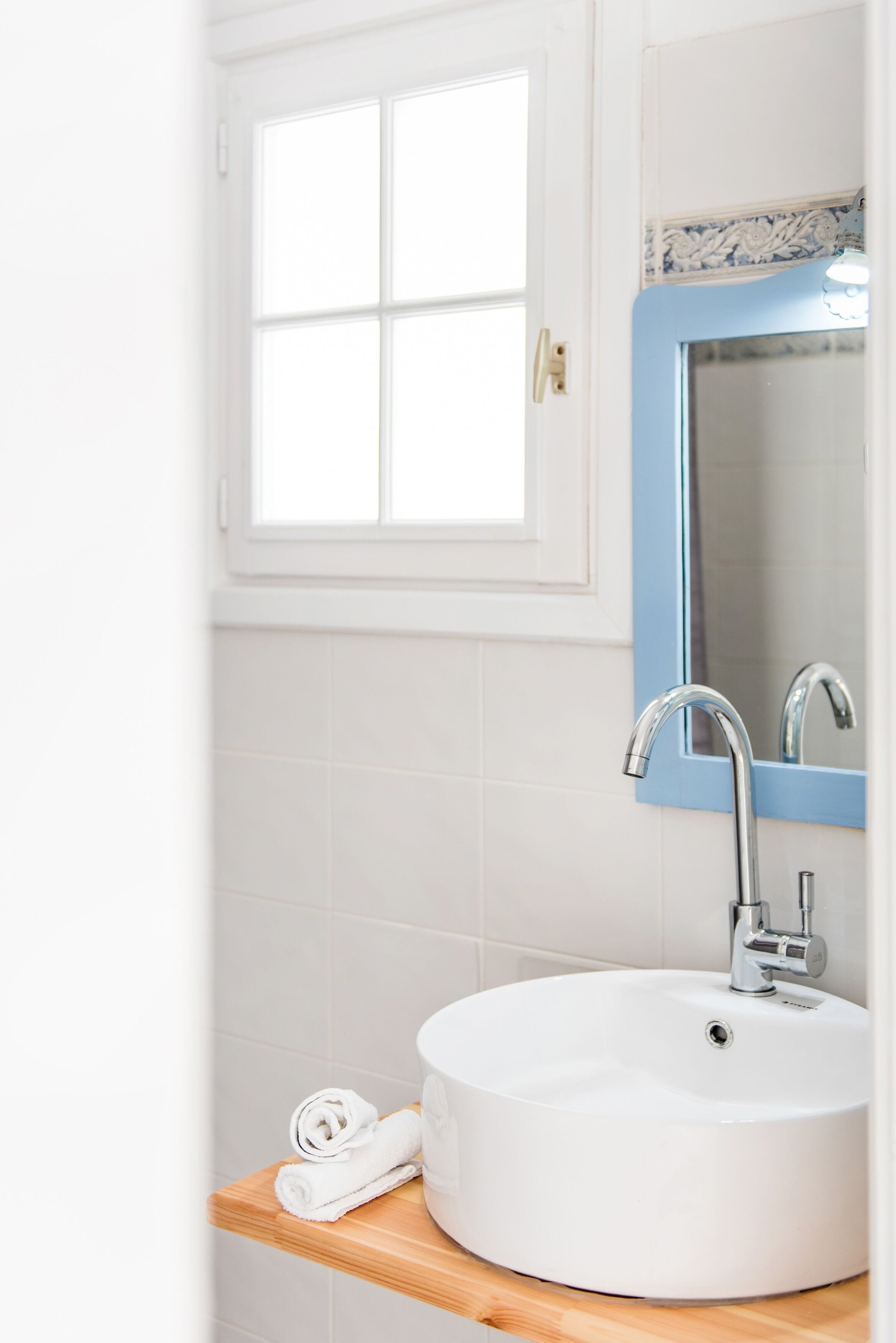 Une salle de bain avec un lavabo blanc, un miroir et une fenêtre située au-dessus du lavabo.