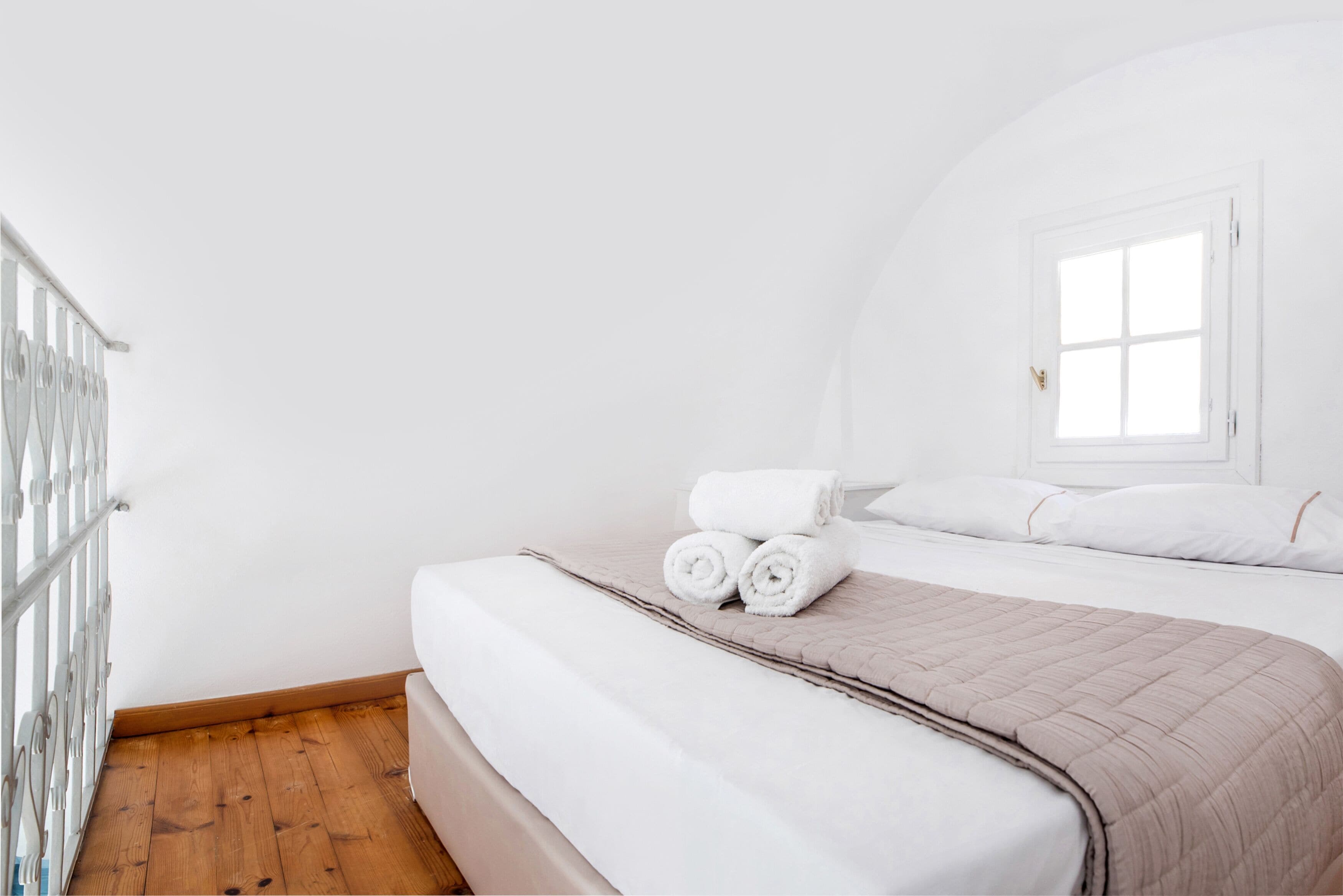 A neatly made bed with rolled towels, a window with white curtains, and a wooden floor.