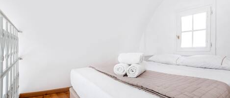 A neatly made bed with rolled towels, a window with white curtains, and a wooden floor.