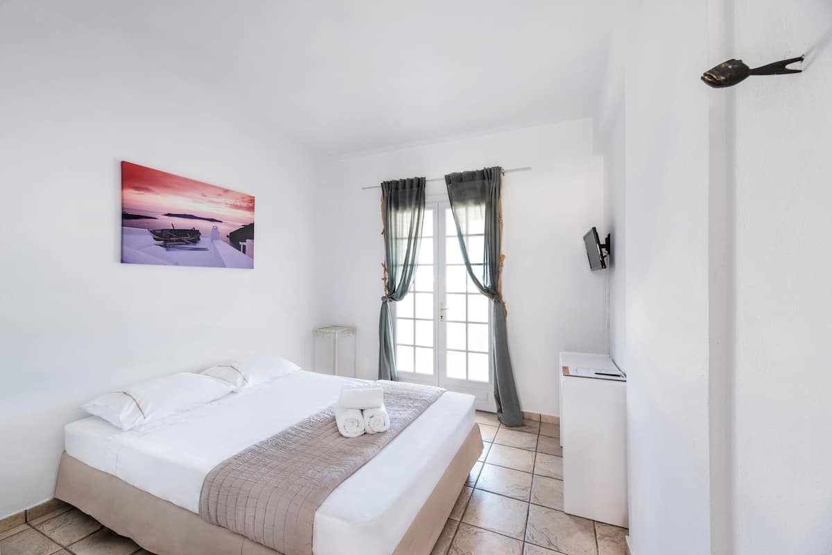 A neatly made bed with white linens and a beige blanket, a small bedside table, a wall-mounted TV, and a window with curtains.