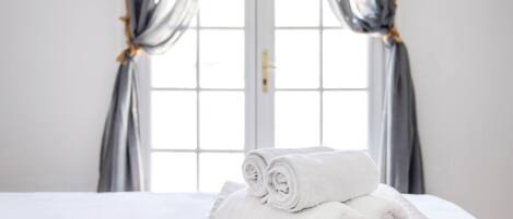 A neatly made bed with rolled white towels, a window with curtains, and a door in the background.