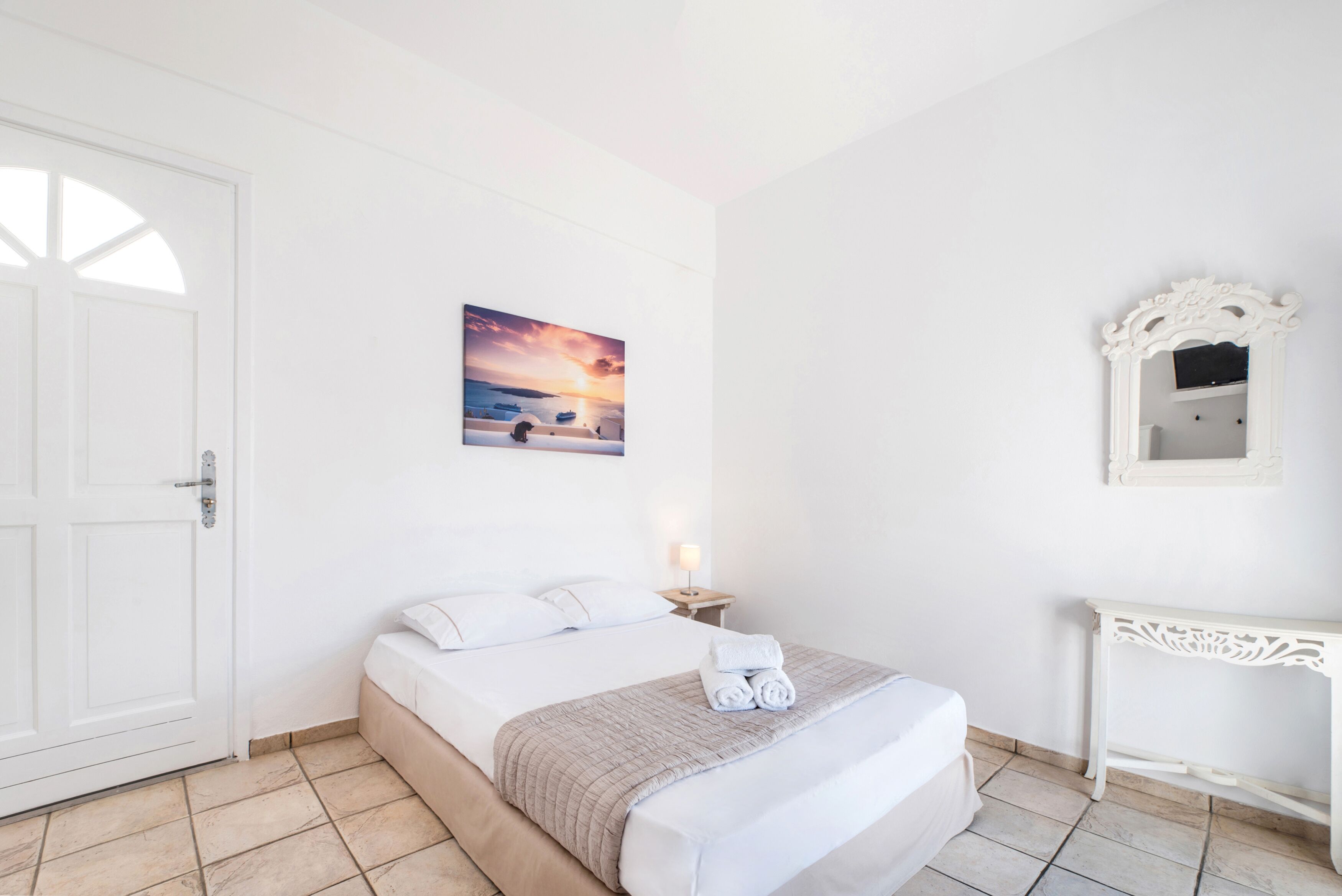 Una habitación de hotel con una cama, un cuadro de un atardecer, una puerta blanca y una mesita consola blanca.