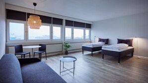 Ein modernes Hotelzimmer mit einer Couch, zwei Betten, einem kleinen Tisch und großen Fenstern.
