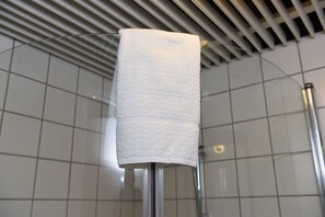 Une serviette blanche est suspendue à un porte-serviettes en métal dans une salle de bain dont les murs sont recouverts de carreaux et dont le plafond est constitué de lattes horizontales.