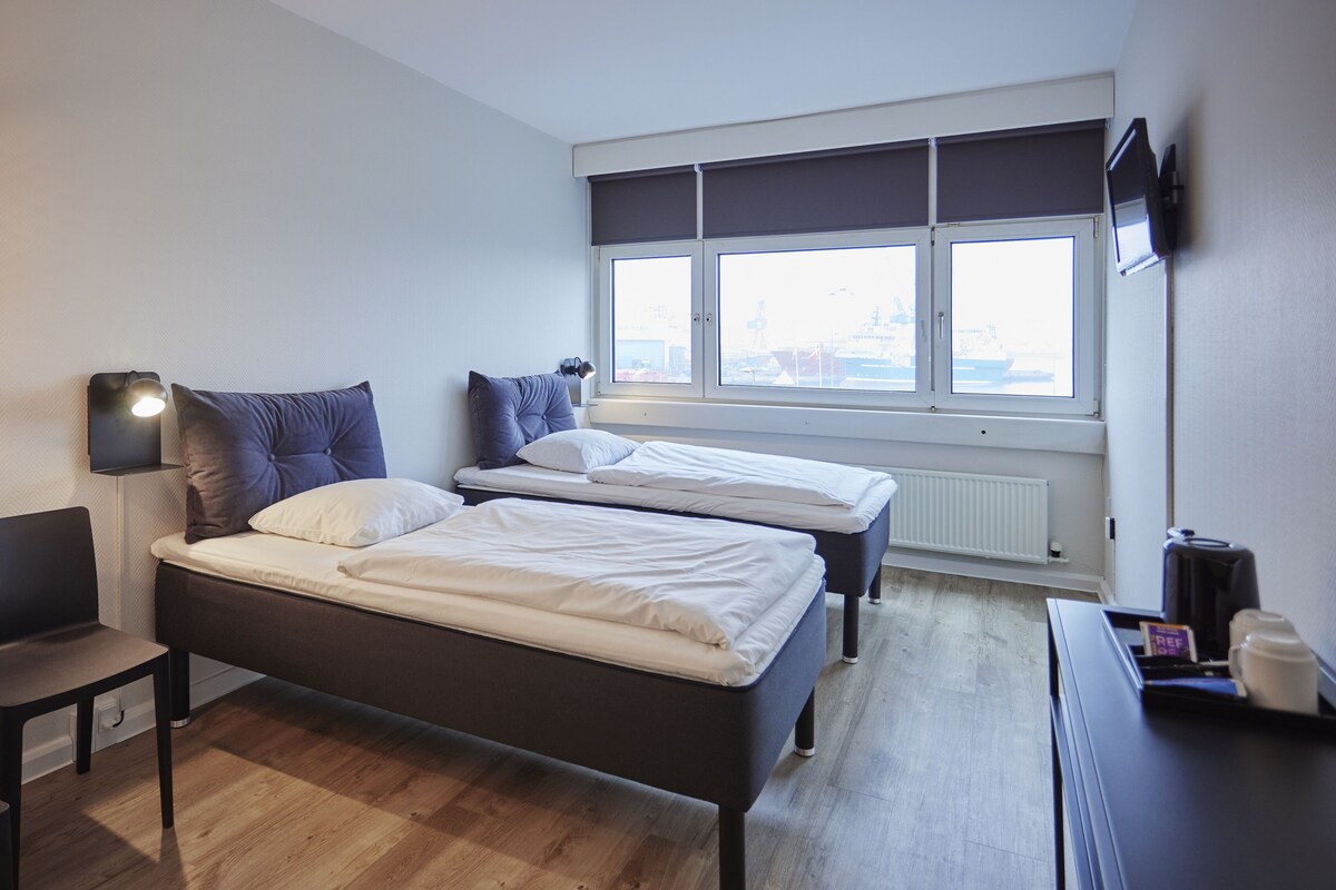 Une chambre d’hôtel moderne, dotée de deux lits, d’un bureau, d’une chaise, d’une lampe et offrant une vue sur la ville grâce à de grandes fenêtres.