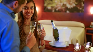 A couple toasting with champagne glasses at a table with a bottle of champagne, a candle, and a framed picture on the wall.