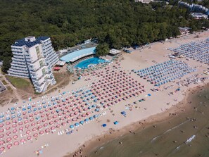 An aerial view of a beach with numerous umbrellas and sun loungers, a swimming pool, and a large hotel building with multiple floors. - Hotel Boryana (Kranevo)