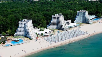 An aerial view of a coastal hotel with multiple high-rise buildings, a large sandy beach, and a swimming pool area.