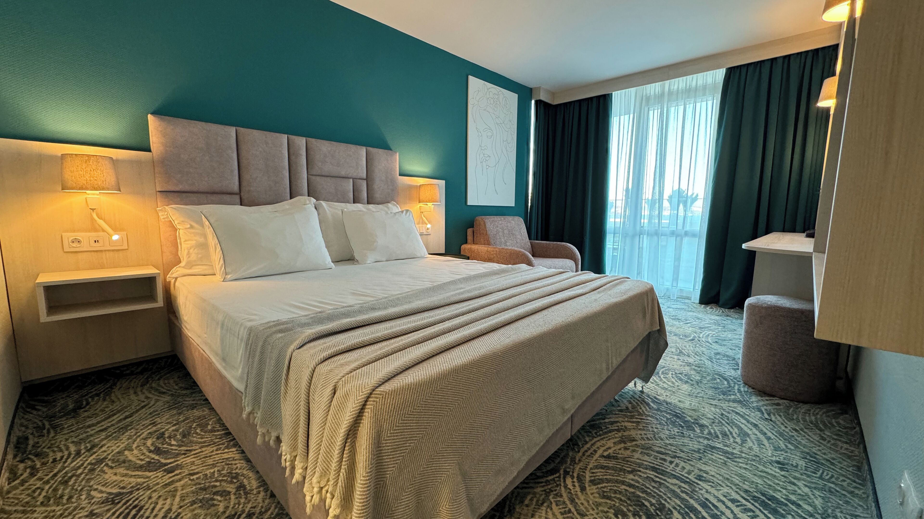 A hotel room with a large bed, a nightstand, a chair, a desk, and a view of the outdoors through a window.