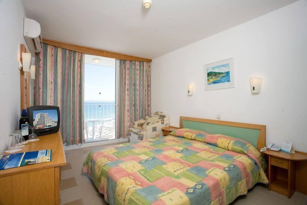 Habitación de hotel con cama, escritorio con televisor, ventana con cortinas y un cuadro en la pared.