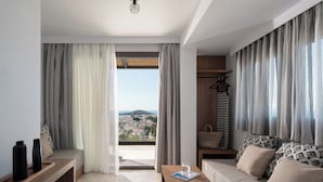 A modern hotel room with a balcony, a sofa with patterned cushions, a wooden side table, and a view of a cityscape. - Poseidon Villas (Skiathos)