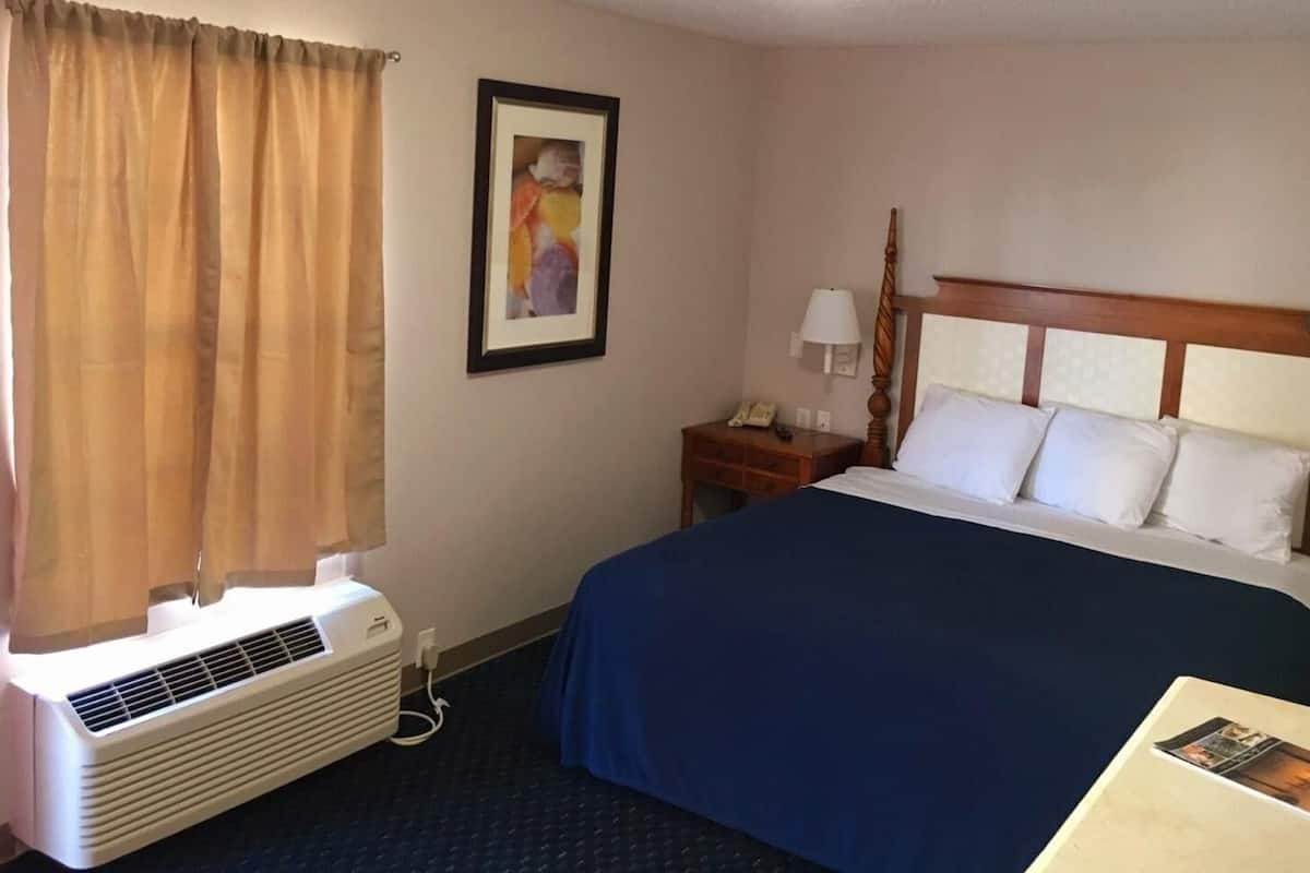 A hotel room with a bed, a nightstand, a lamp, a framed picture, and an air conditioning unit.