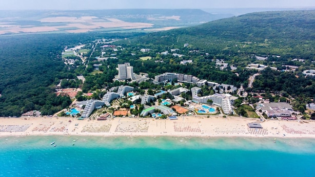 An aerial view of a coastal resort with multiple hotel buildings, a beach, and surrounding greenery. - Hotel Flamingo (Albena)