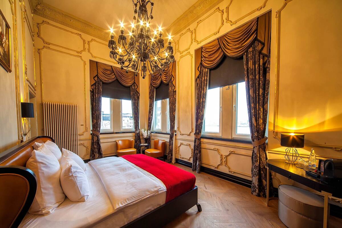 Una lujosa habitación de hotel con una cama grande, un candelabro, cortinas ornamentadas y vistas a la ciudad.