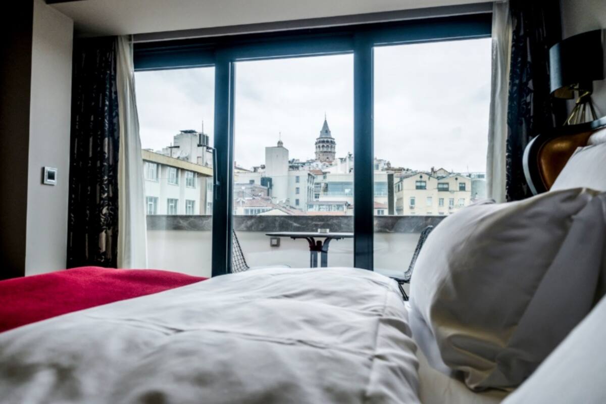 Habitación de hotel con cama, una colcha roja y vistas de un paisaje urbano a través de ventanas grandes.