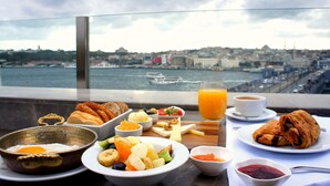 A breakfast table with various foods and a glass of orange juice, overlooking a cityscape with a body of water and buildings. - Nordstern Hotel Galata (Istanbul)
