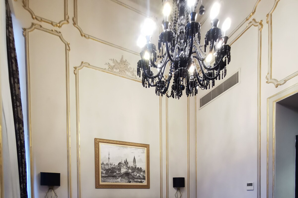 A hotel room with a large bed, a chandelier, a framed cityscape picture, and a bedside table with a lamp.