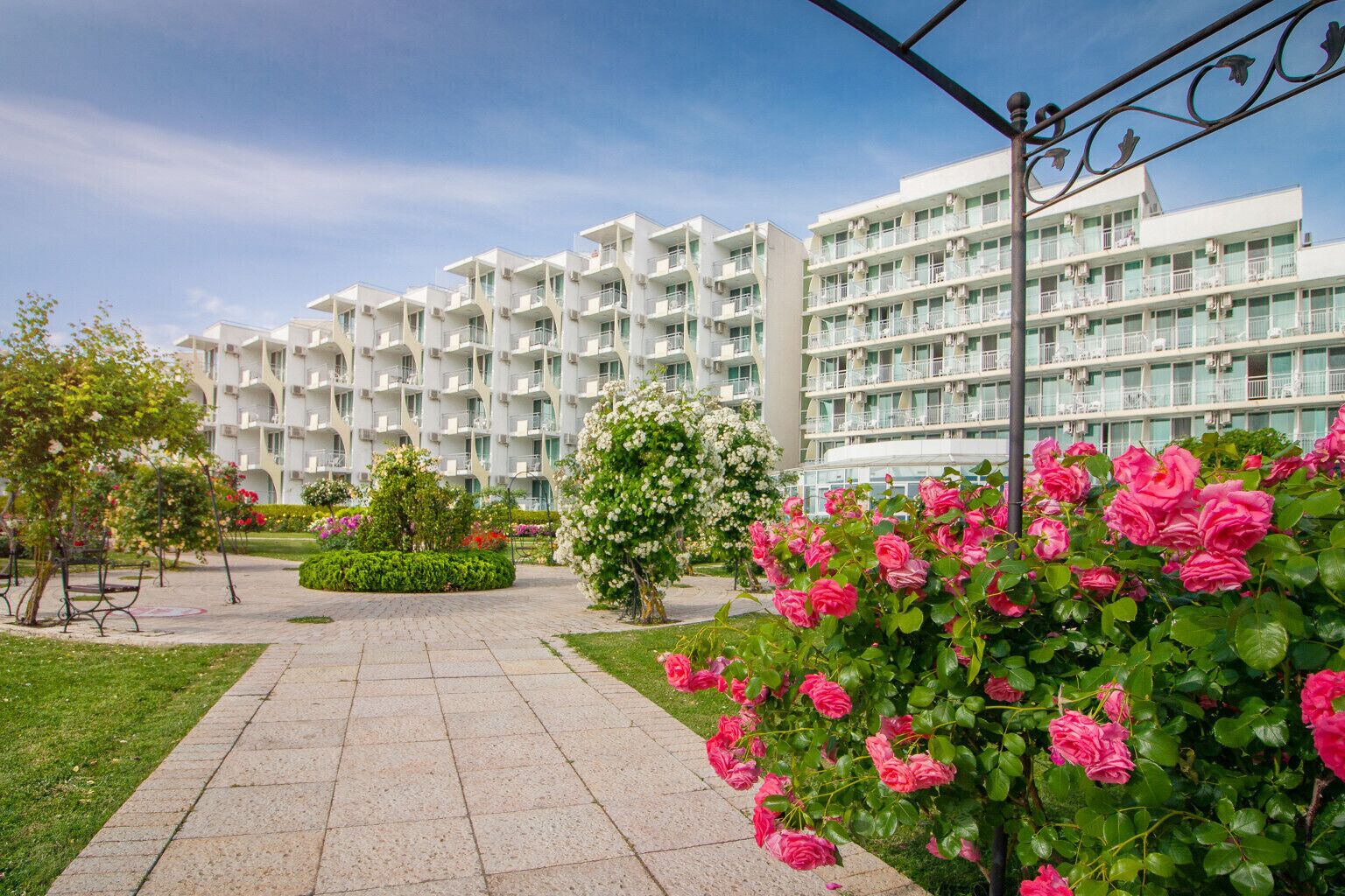 A modern hotel with multiple floors, a paved walkway, and well-maintained gardens with pink roses.