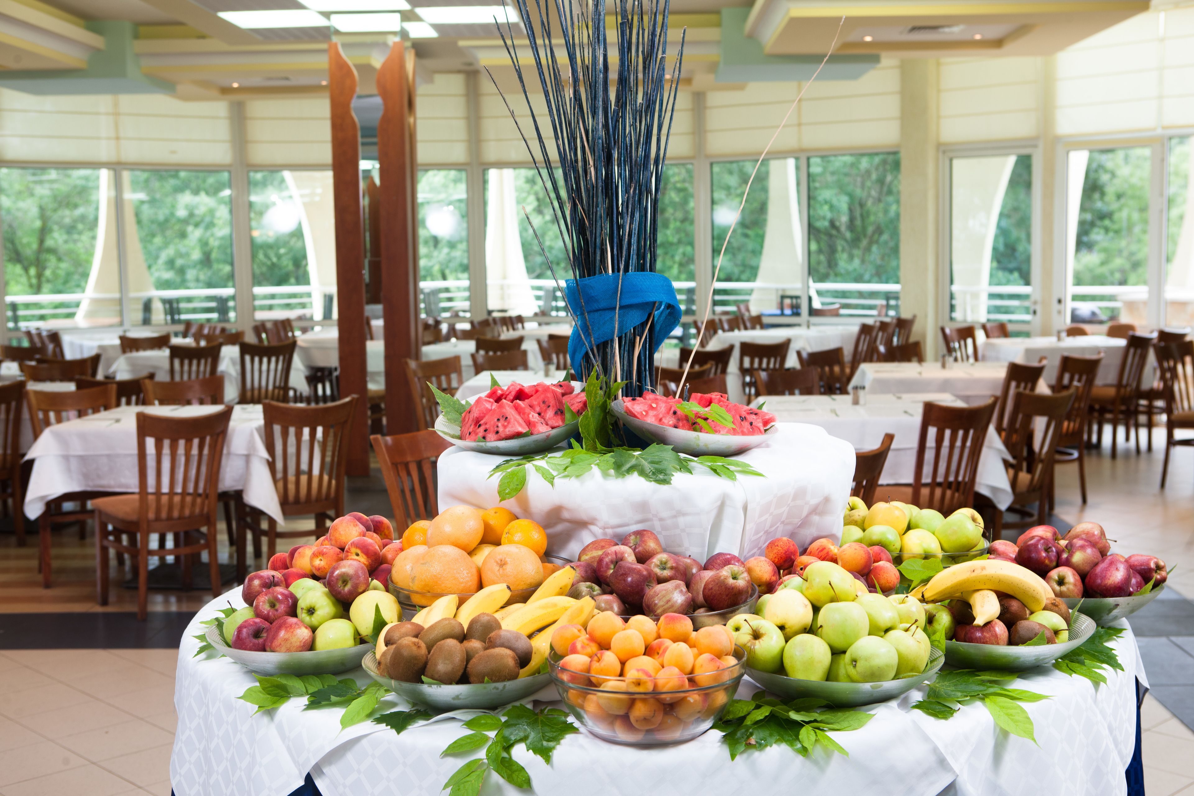 A buffet table with various fruits and vegetables, surrounded by dining tables and chairs in a spacious room with large windows.