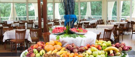 A buffet table with various fruits and vegetables, surrounded by dining tables and chairs in a spacious room with large windows.