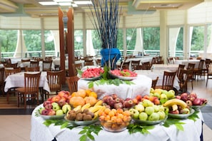 A buffet table with various fruits and vegetables, surrounded by dining tables and chairs in a spacious room with large windows.