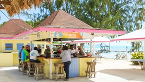 A beachside bar with wooden stools and a pink awning. - Anegada Reef Hotel (Setting Point)