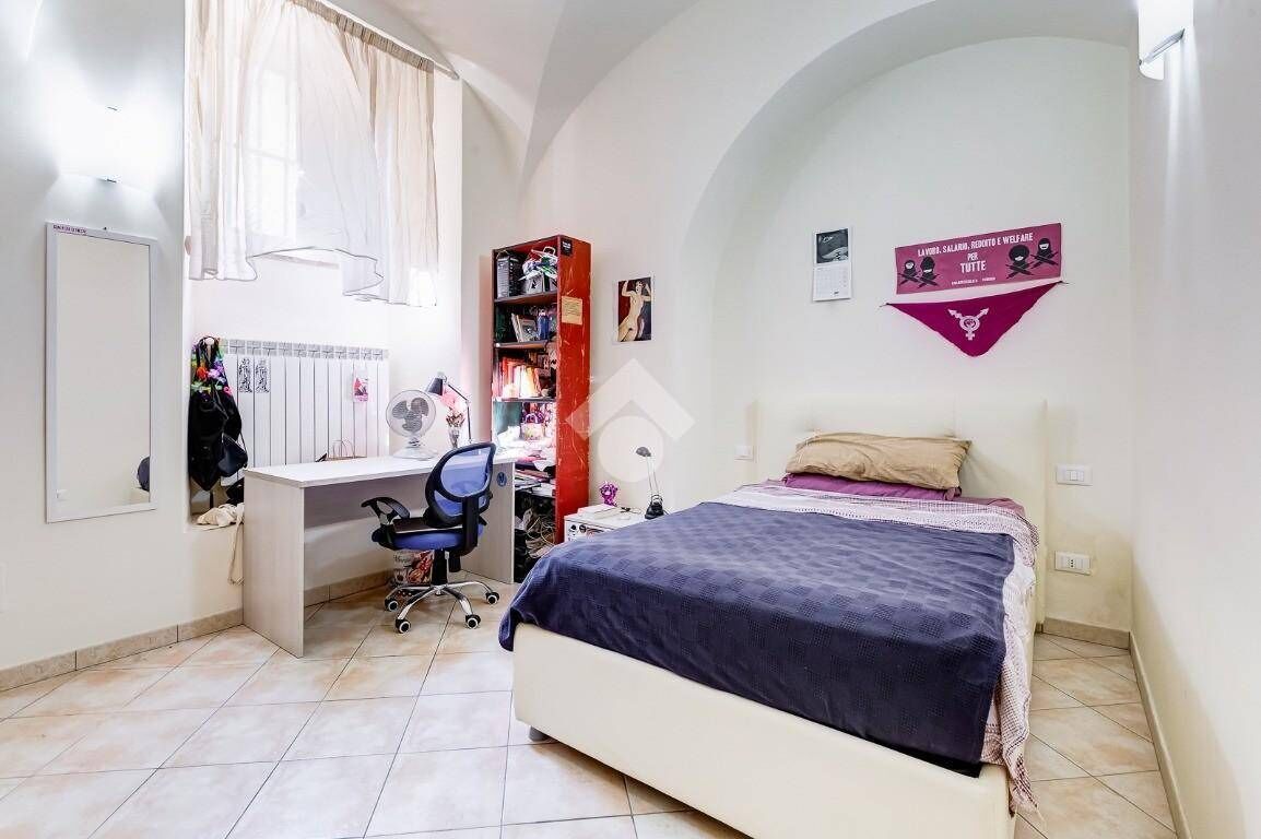 A bedroom with a bed, a desk, and a chair. There is a window with curtains, a bookshelf, and a wall-mounted fan.