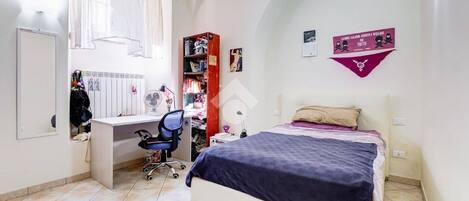 A bedroom with a bed, a desk, and a chair. There is a window with curtains, a bookshelf, and a wall-mounted fan.