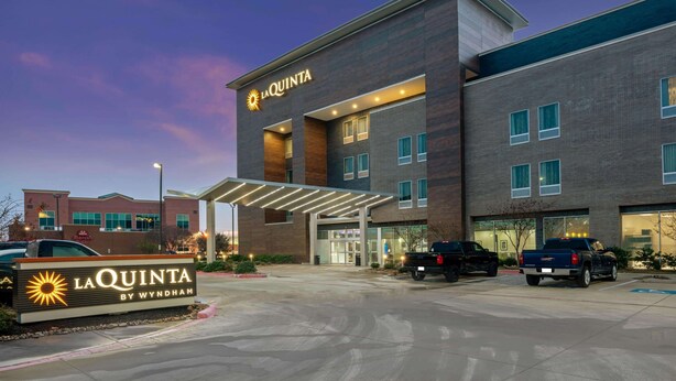 A La Quinta hotel with a modern design, featuring a prominent entrance and a parking area with several vehicles.