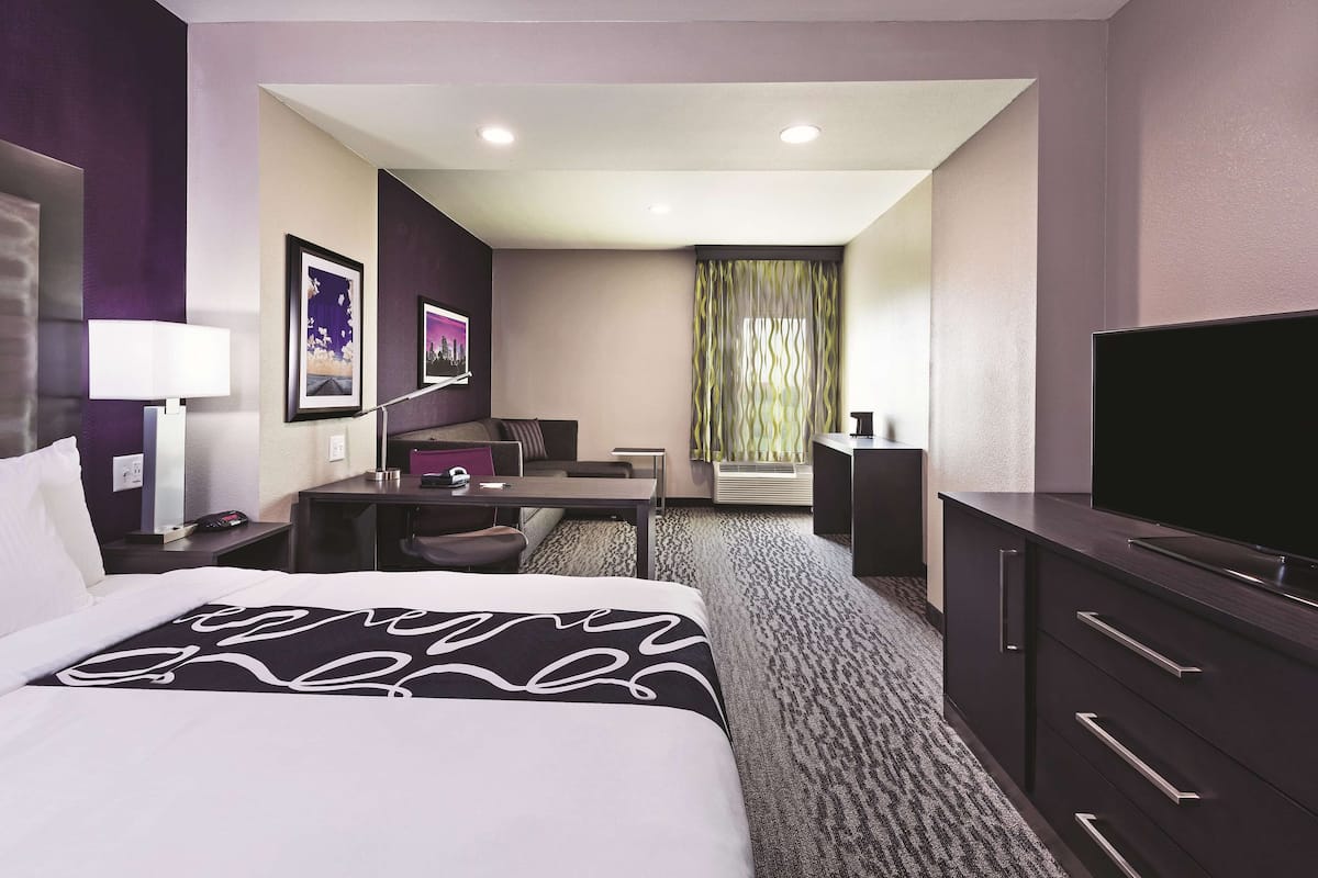 Habitación de hotel con cama, escritorio, silla, televisor y ventana con cortinas.