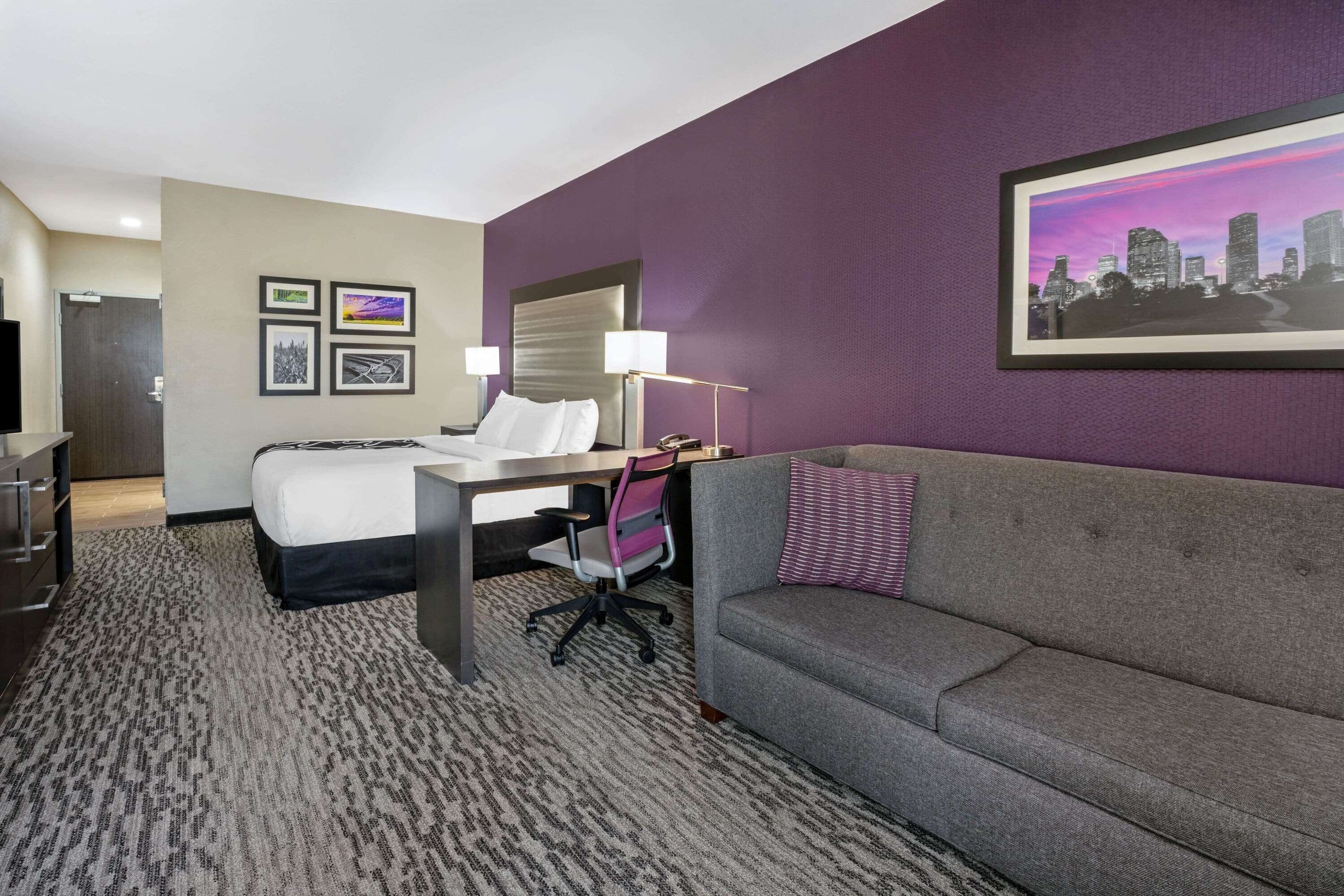 Una habitación de hotel con cama, escritorio, sofá gris y un cuadro de paisaje urbano en la pared.