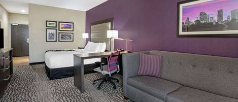 A hotel room with a bed, a desk, a grey sofa, and a cityscape painting on the wall.