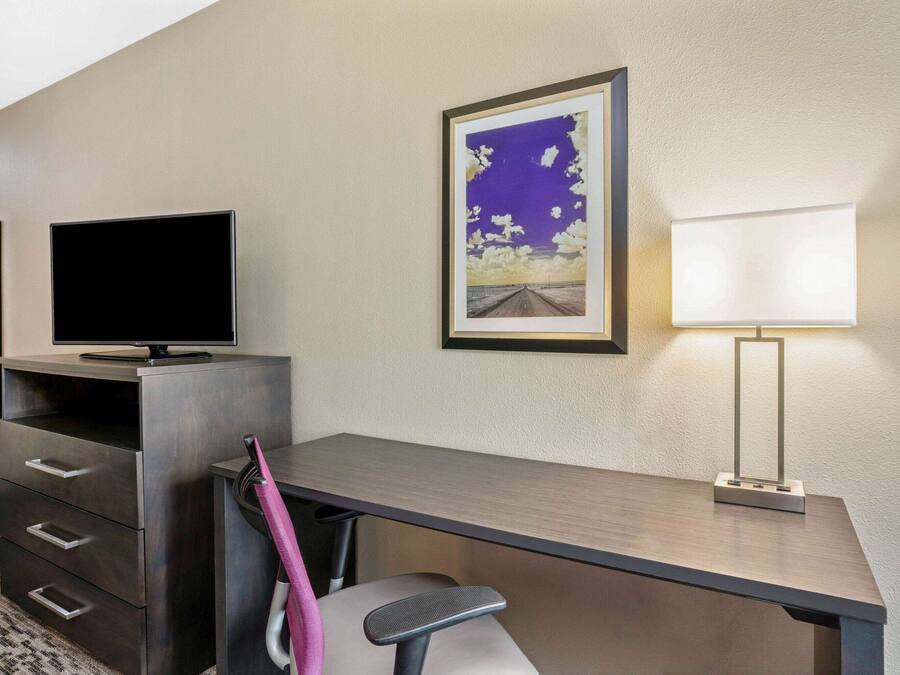 A hotel room with a desk, a flat-screen TV, a framed picture, and a bedside lamp.