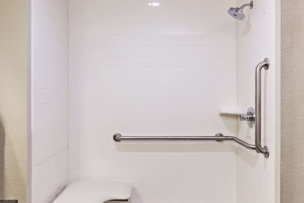 Ducha a pie con paredes de azulejos blancos, un asiento y una barra de apoyo.