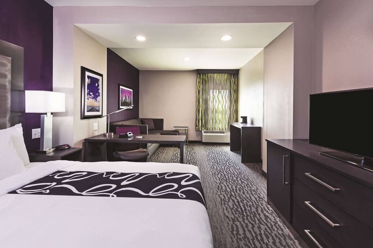 Una habitación de hotel con cama, escritorio, silla, televisor y ventana con cortinas.