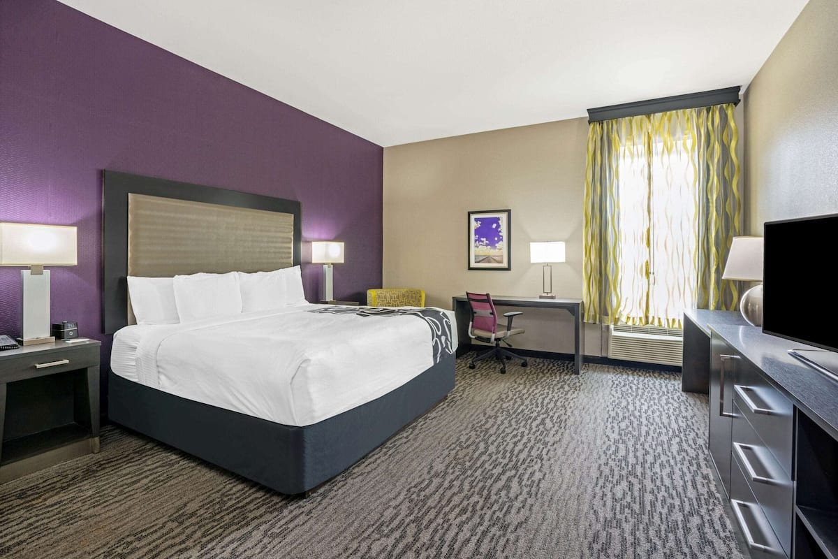 Habitación de hotel con cama, escritorio, silla, televisor y ventana con cortinas.