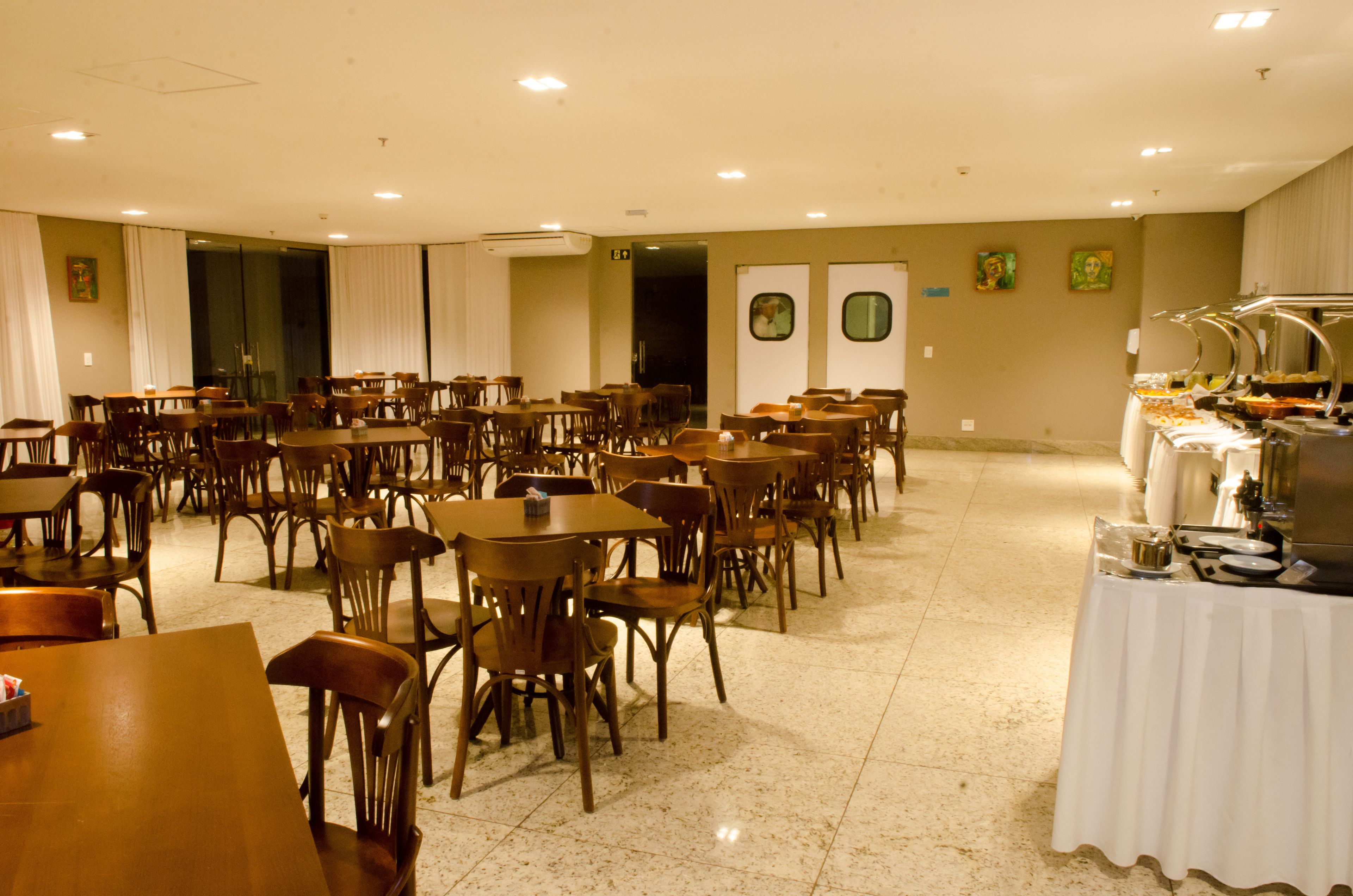 Uma ampla área de refeições com várias mesas e cadeiras, um buffet de um lado e uma grande janela que proporciona luz natural.