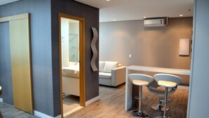 Quarto de hotel moderno com porta de madeira, banheiro com chuveiro de vidro, sofá branco e dois bancos altos.