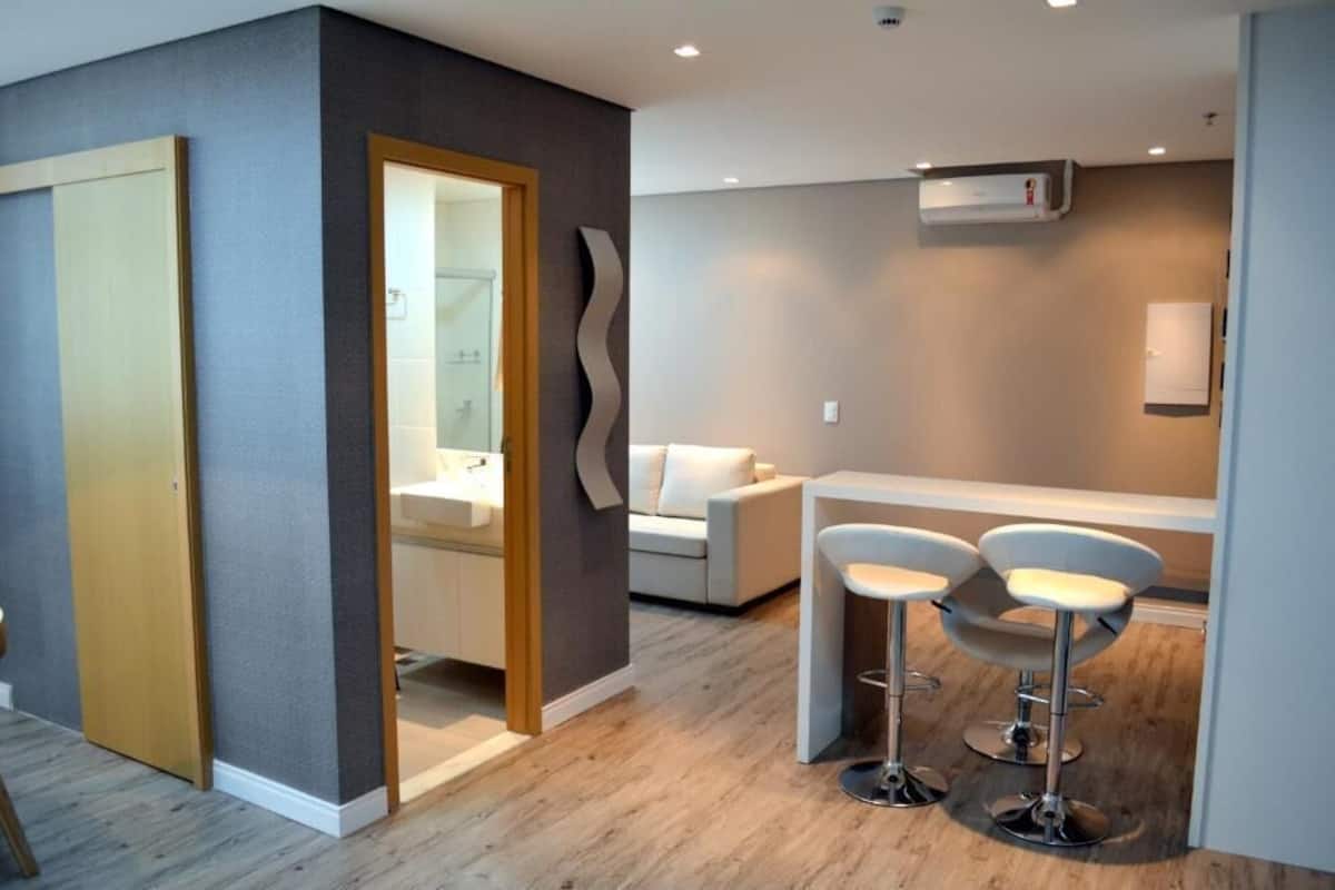 A modern hotel room with a wooden door, a bathroom with a glass shower, a white sofa, and two bar stools.