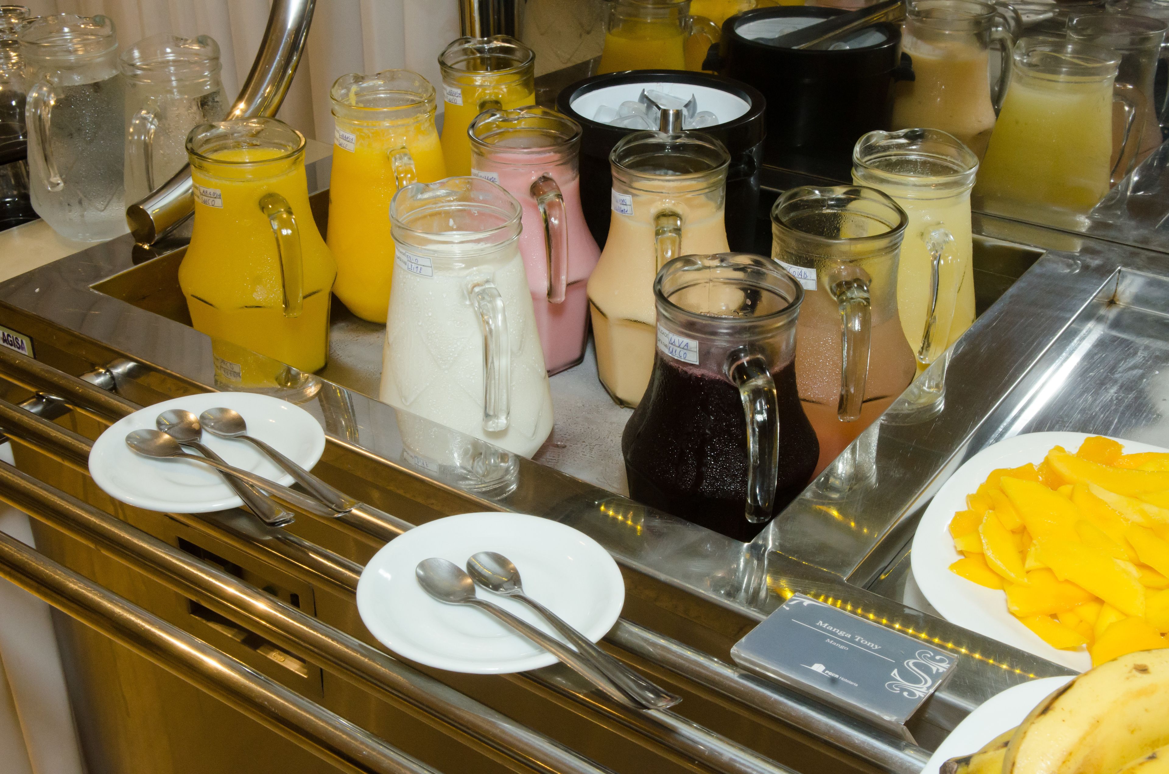 Uma área de buffet com diversas bebidas em jarras e copos, pratos com talheres e um prato de fruta fatiada.