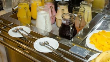 Uma área de buffet com diversas bebidas em jarras e copos, pratos com talheres e um prato de fruta fatiada.