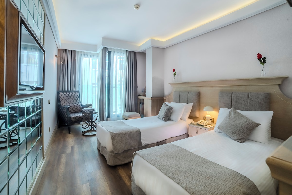 A hotel room with two beds, a desk, a chair, a nightstand with a lamp, and a window with curtains.